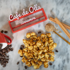 Popcorn, coffee beans, cinnamon sticks, and brown sugar on a marble surface with a red cup and a "Cafe de Olla" sign.