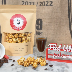 Coffee popcorn bag with scattered beans, a coffee cup, and a glass next to a "Flat White" sign on a burlap background.