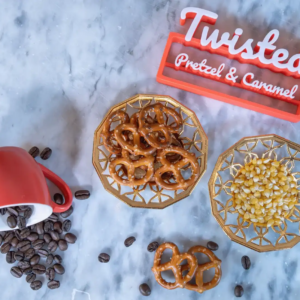 Pretzels and popcorn on plates, red cup with coffee beans, and "Twisted Pretzel & Caramel" sign on marble surface.