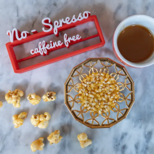 Caffeine-free coffee in red cup, popcorn kernels in decorative dish, caramel popcorn scattered, and "No-S presso" sign.