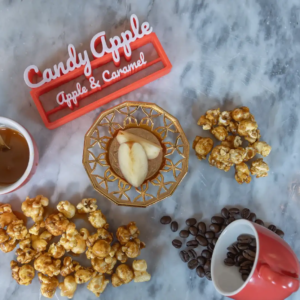 Caramel popcorn, apple slices, coffee beans, and a caramel-filled mug on marble with a "Candy Apple" sign.
